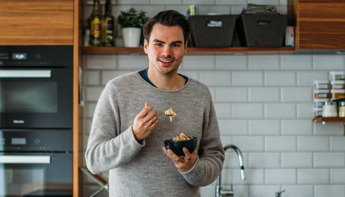 Unser Redakteur Florian in unserer Küche. Er trägt einen grauen Pullover und lacht fröhlich in die Kamera während er (wie immer) etwas isst.