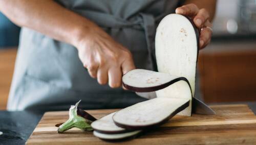 Darf man Auberginen roh essen? Diese Frage beantwortet dir unser Redakteur Florian in diesem Artikel. Auf dem Bild ist eine Aubergine zu sehen, die gerade auf einem Holzbrett in Scheiben geschnitten wird.