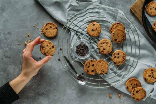 Chocolate-Chip-Cookies in Hand und auf Gitter mit Schokodrops vor hellem Hintergrund.