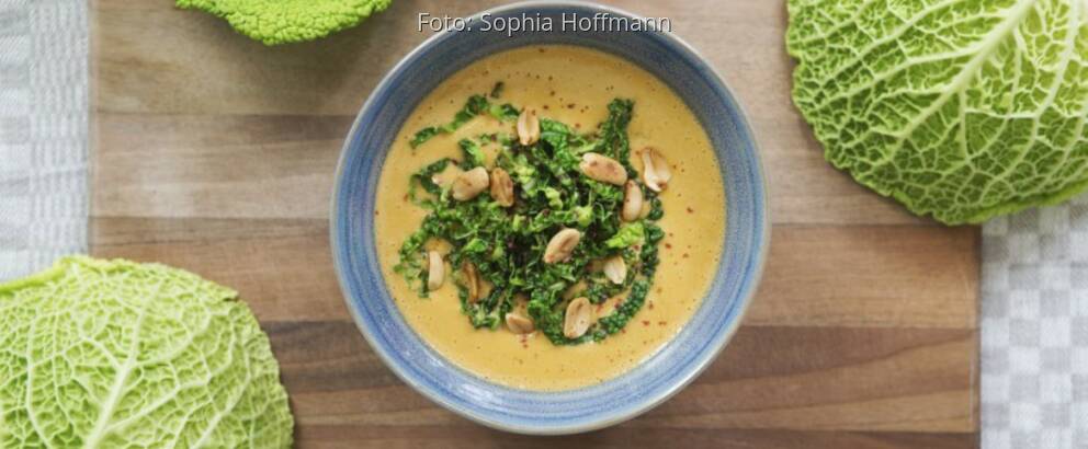 Wirsing-Erdnuss-Suppe in grauer Schale auf Holzbrett mit Wirsingblättern dekoriert