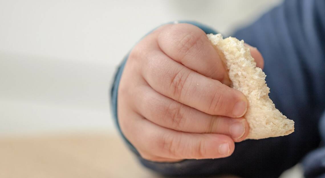 Kinderhand mit einem Stück Brot in der Hand, Nahaufnahme vor hellem Hintergrund. Kind trägt dunkelblauen Pullover. 
