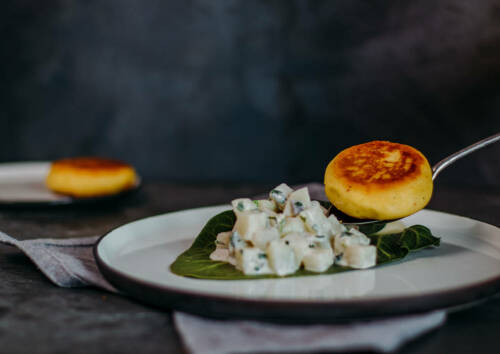 Weißer Teller auf dem ein Kohlrabiblatt mit Kohlrabigemüse zu sehen ist. Außerdem ein goldgelbes Kartoffelküchlein auf einem Löffel vor dunkelgrauem Hintergrund. Von vorne fotografiert.