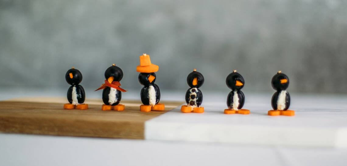 Pinguine aus Oliven mit Karotten und Dip deiner Wahl. 6 Pinguine in einer Reihe auf einem Küchenbrett vor grauem Hintergrund. Von vorne fotografiert.