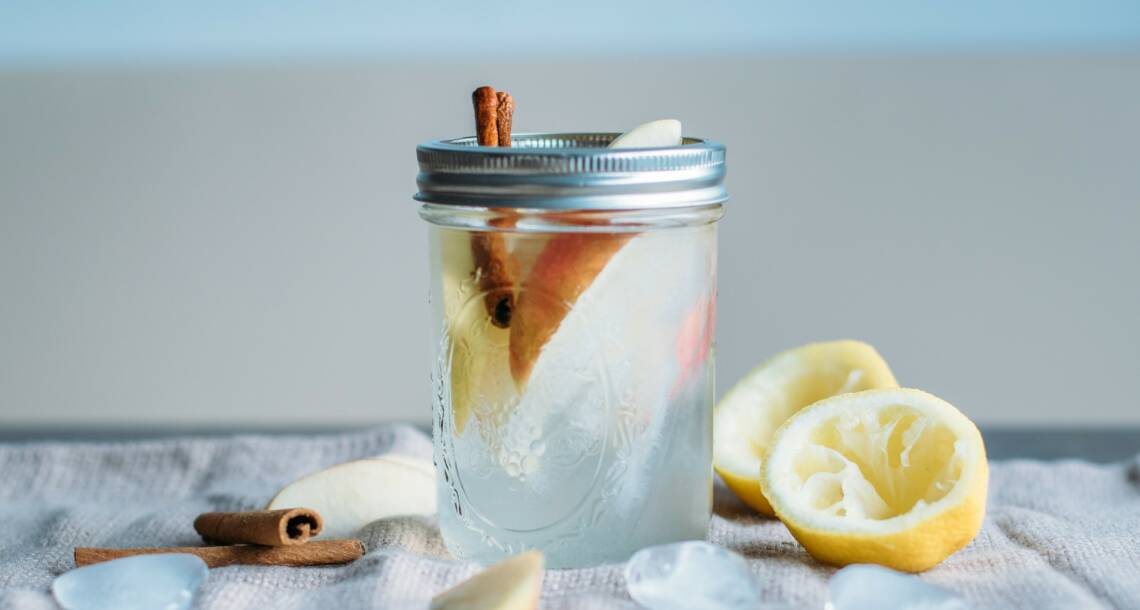 Glas gefüllt mit Wasser, Zimtstange und Apfelschnitzen Vor hellem Hintergrund, daneben Zitronen und Zimtstangen. Von der Seite fotografiert.