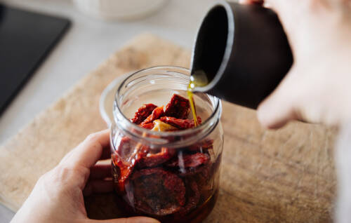 getrocknete Tomaten einlegen in Olivenöl und Schraubglas mit Händen vor hellem Hintergrund.