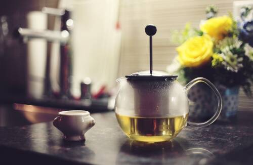 Einer von zwei natürlichen Stoffwechsel-Boostern, denen wissenschaftlich eine Wirkung nachgewiesen wurde, ist grüner Tee. Hier ist eine Kanne grüner Tee zu sehen, im Hintergrund eine Küche und Blumen.