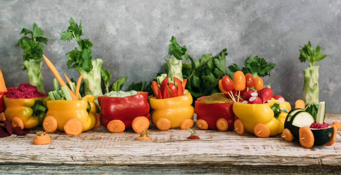 Snack Zug aus Paprika, gefüllt mit Gemüse-Dips, Gurkensticks, Tomaten, Karotten und Hummus. Vor grauem Hintergrund, von der Seite fotografiert.