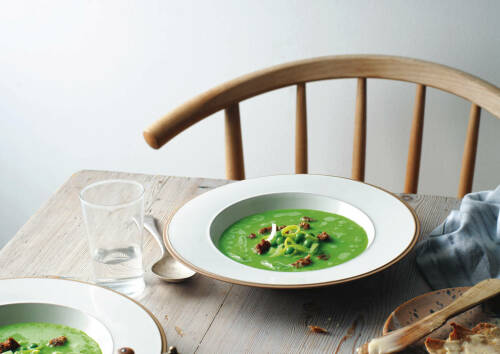Grüne Erbsensupper in weißem Teller auf einem Holztisch, dekoriert mit Lauchscheiben