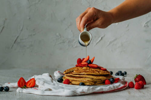 Leinsamen Pancakes auf Teller mit Beeren und Hand mit Ahornsirup vor hellem Hintergrund.