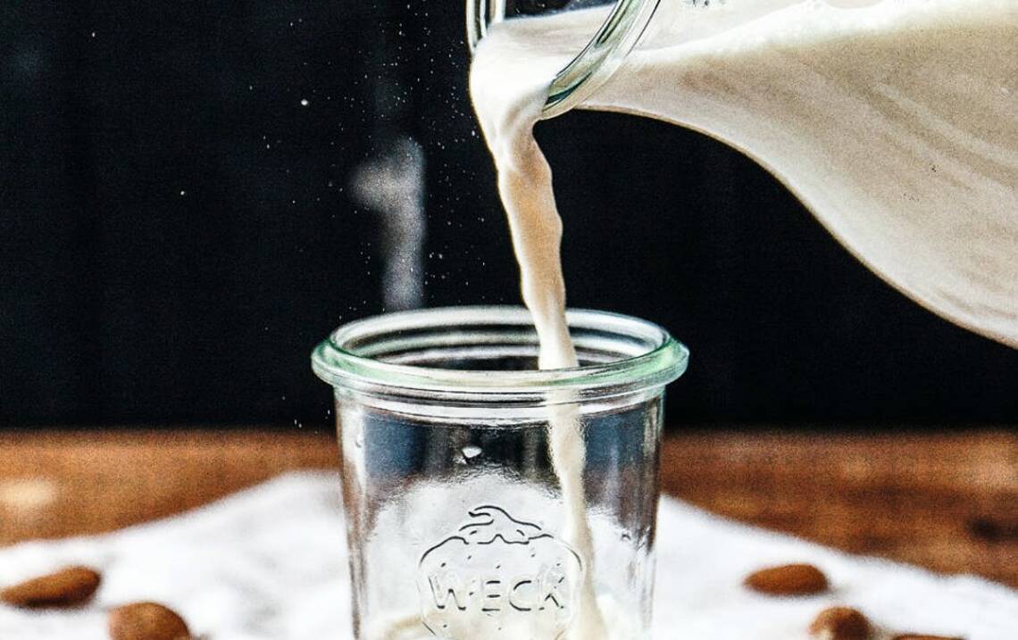 Aus einer Karaffe wird Mandelmilch in ein Glas gegossen, der Hintergrund ist schwarz. Von der Seite fotografiert.