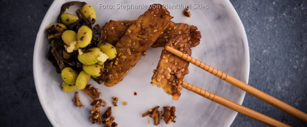Weißer Teller mit mariniertem Tempeh und Edamame, dazu Essstäbchen auf dunkler Arbeitsplatte