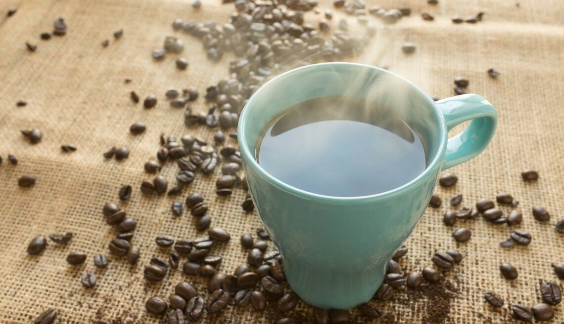 Blaue Tasse gefüllt mit dampfendem Kaffee auf einer Bastmatte, daneben lose Kaffeebohnen, von oben fotografiert. 