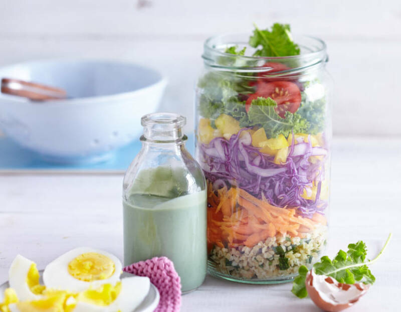 Regenbogensalat in Glas neben Dressing und gekochten Eiern vor hellem Hintergrund.