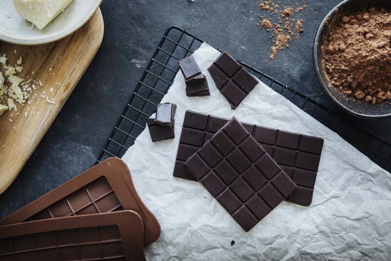 Ausgekühlte selbstgemachte Schokolade auf Gitter neben Kakao und Kakaobutter vor dunklem Hintergrund.