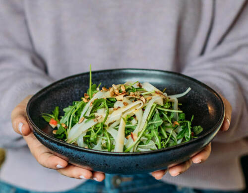 Schwarzwurzel-Salat in dunklem Teller mit zwei Händen vor hellem Hintergrund.