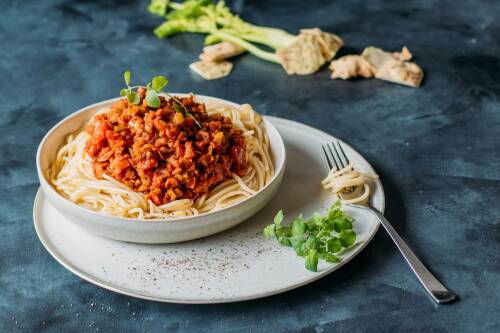 Spaghetti mit Selleriebolognese auf weißem Teller neben Sellerie und Gabel vor dunklem Hintergrund.