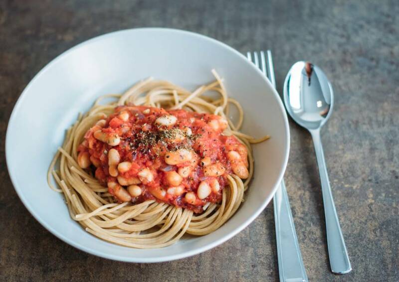 Portion Spaghetti mit Tomaten-Sauce und weißen Bohnen in einer weißen Schüssel, daneben Besteck.