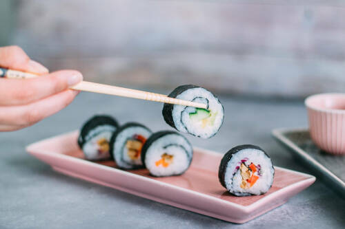 Vegetarisches Sushi auf Teller mit Hand und Stäbchen vor hellem Hintergrund.