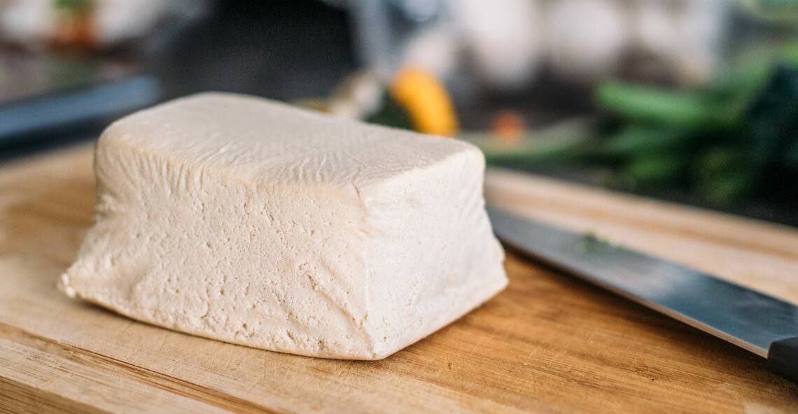 Roher, unverarbeiteter  Tofu auf einem Schneidebrett. Weiß-gräuliche Farbe. Daneben ein Messer, von der Seite fotografiert.