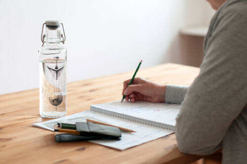 Eine transparente Trinkflasche steht auf einem Schreibtisch, an dem jemand sitzt und auf einen Block schreibt.