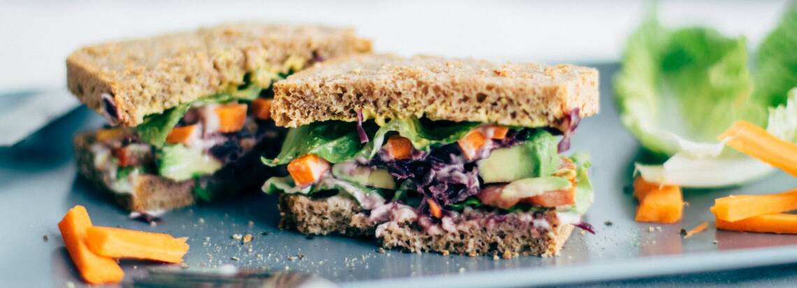Alles, was du über vegane Ernährung wissen musst, erfährst du in diesem Artikel. Außerdem gibt es viele leckere Rezepttipps, wie dieses Avocado-Veggie Sandwich. Sandwich auf einer Platte mit Salat und Karotten.