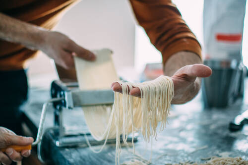 Bei der Herstellung von veganer Pasta kann eine Nudelmaschine verwendet werden. Auf dem Bild kann man die Herstellung von Spaghetti sehen, die gerade durch die Nudelmaschine gedreht werden.