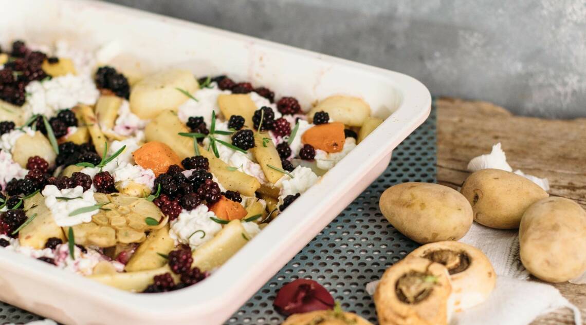 Auflaufform gefüllt mit Ofengemüse aus Karotten und Pastinaken, darüber Hüttenkäse und Brombeeren, der Hintergrund ist hellgrau, von oben fotografiert.